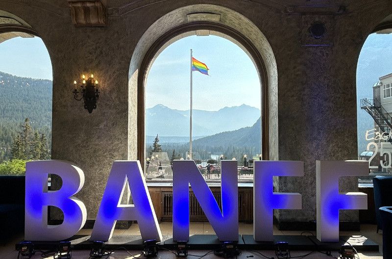 Dans une élégante pièce aux fenêtres cintrées en pierre, de grandes lettres 3D illuminées d'une lumière bleue forment le mot « BANFF ». À travers la fenêtre centrale, un drapeau arc-en-ciel, symbole de la fierté LGBTQ+, flotte fièrement sur un ciel d'un bleu limpide, avec en arrière-plan des montagnes couvertes de pins et une vallée pittoresque.
