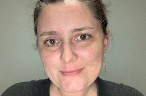 Portrait d'une femme souriante aux cheveux bruns attachés, portant des boucles d'oreilles pendantes en argent et un t-shirt noir, sur fond gris.