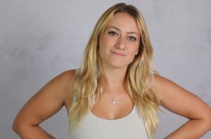 Une femme blonde aux cheveux longs, debout, les mains sur les hanches, vêtue d'un débardeur beige, sur un fond neutre.